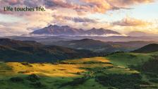 Green expanse and outstretched mountains under a colorful cloudy sunset.